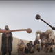 A Tusken clad in black teaches Boba Fett to wield his gaderffii stick as others watch on.